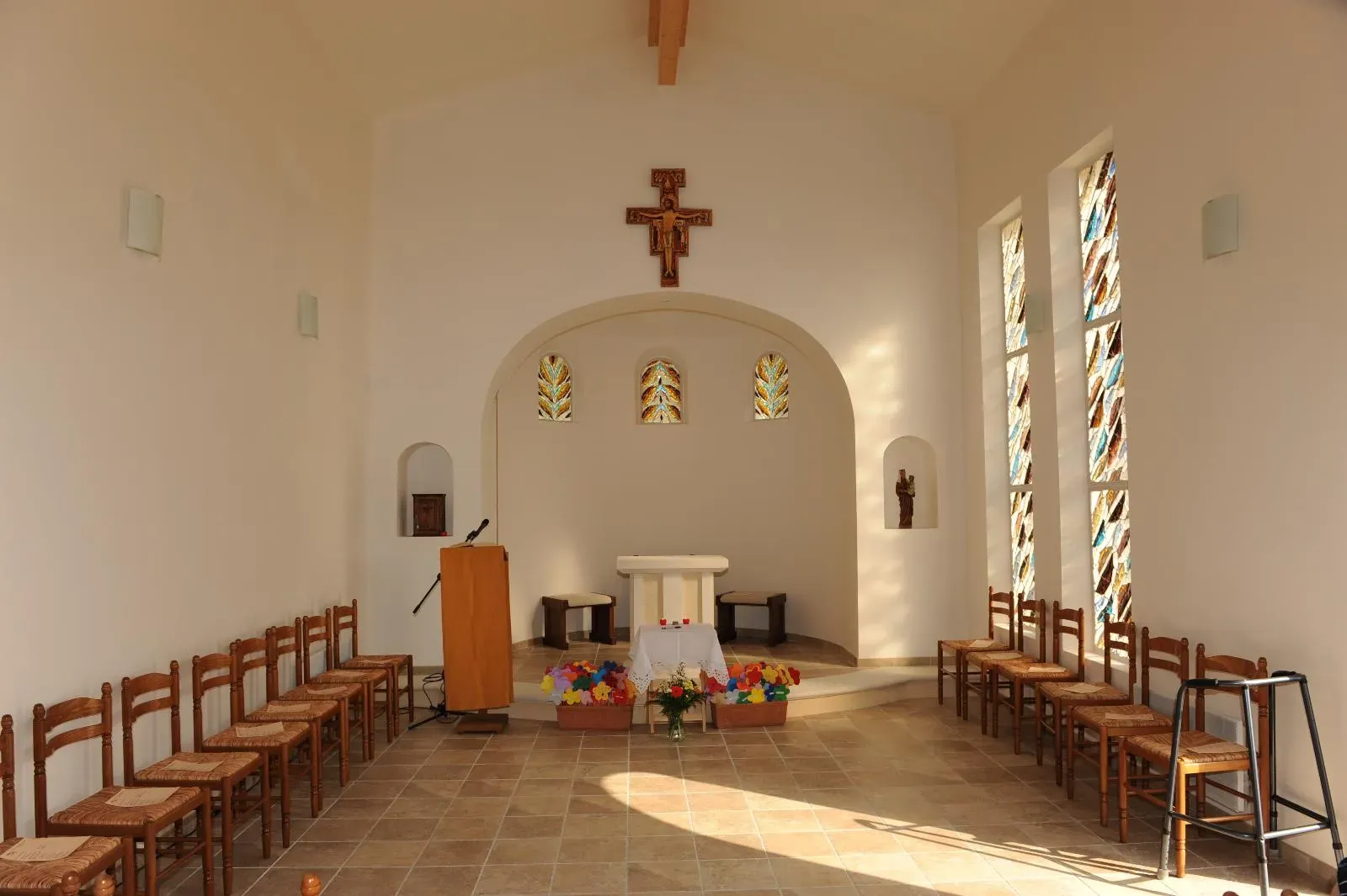Intérieur d’une chapelle aux murs blancs, abside voûtée, vitraux colorés et chaises paillées