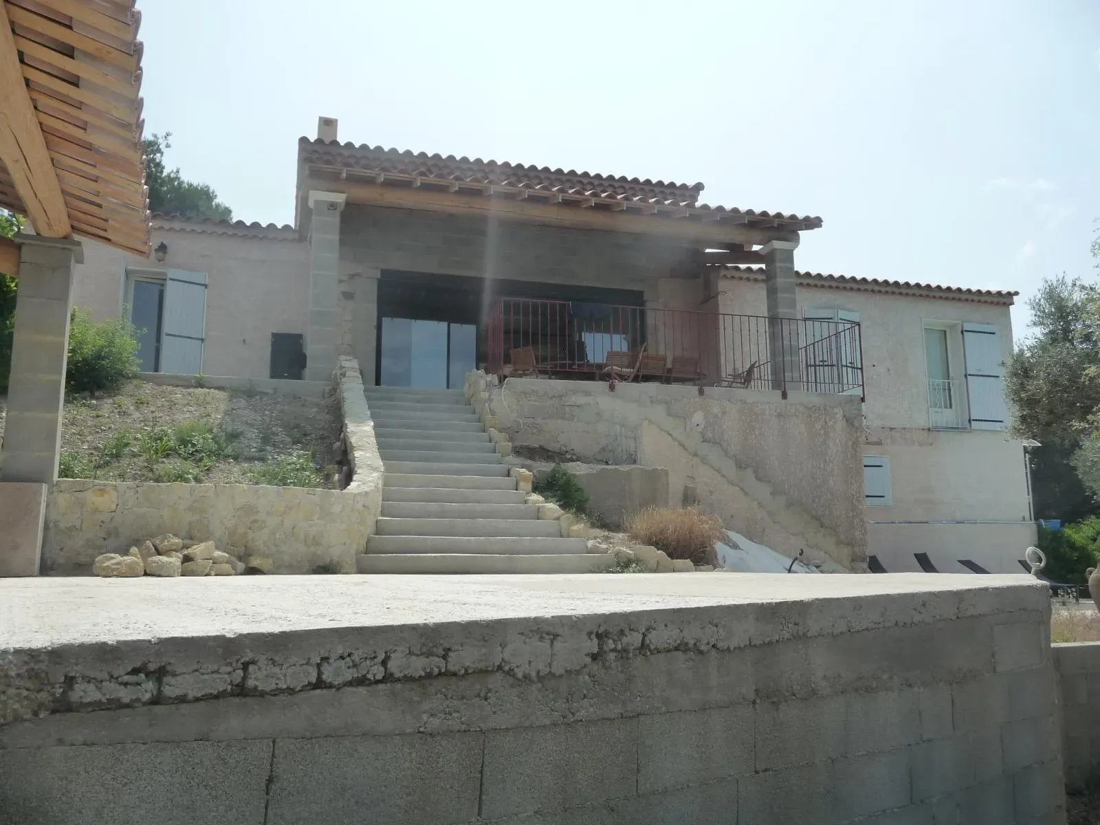 Maison en pente avec escalier extérieur en béton, murets de pierre et terrasse couverte sur piliers