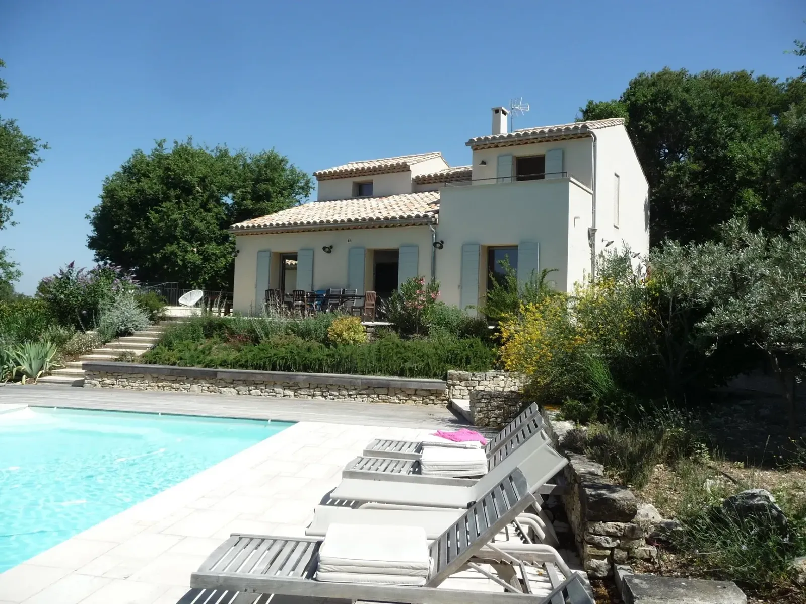 Maison aux façades claires et volets gris-bleu au-dessus d’une piscine, jardin en restanques de pierre sèche et transats