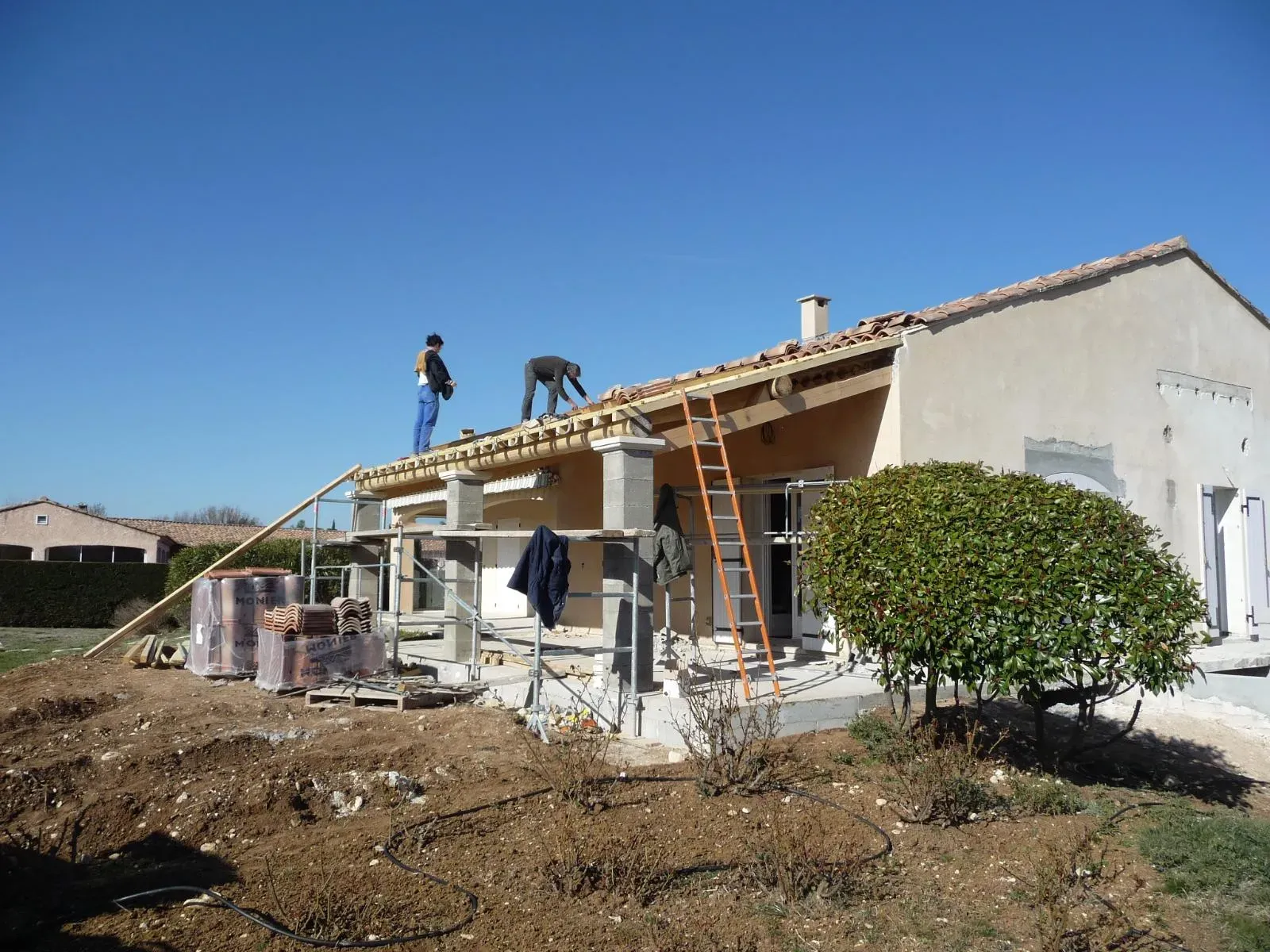 Deux personnes posent des tuiles sur la charpente neuve d’un auvent, échafaudage et palettes de tuiles devant la maison