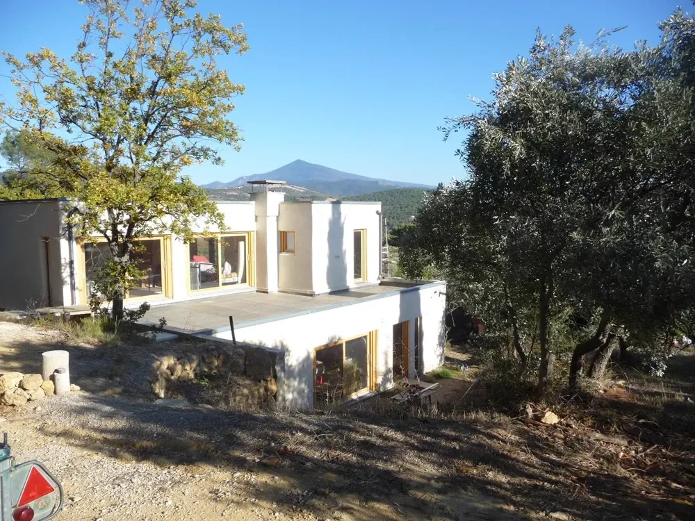 Maison contemporaine aux toits-terrasses parmi les oliviers, relief boisé et sommet à l’horizon